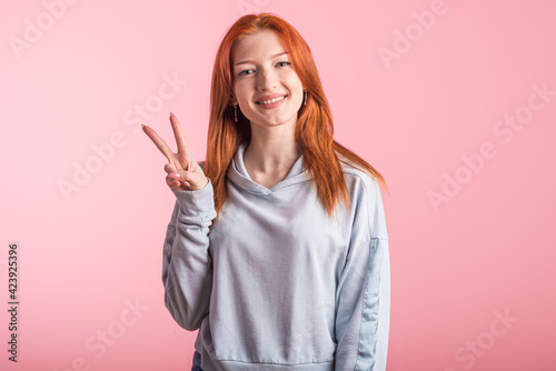 Young redhead girl showing peace gesture in studio on pink background © Павел Костенко
