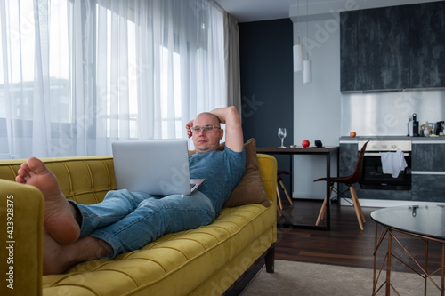 Man in a blue t-shirt lying on the yellow sofa and using laptop. Remote work, work from home. © alurk