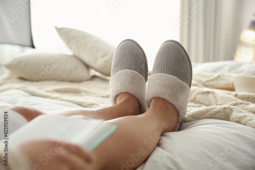 Woman in soft comfortable slippers reading book on bed at home, closeup