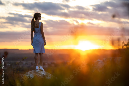A young woman in summer dress standing outdoors enjoying view of bright yellow sunset. © bilanol