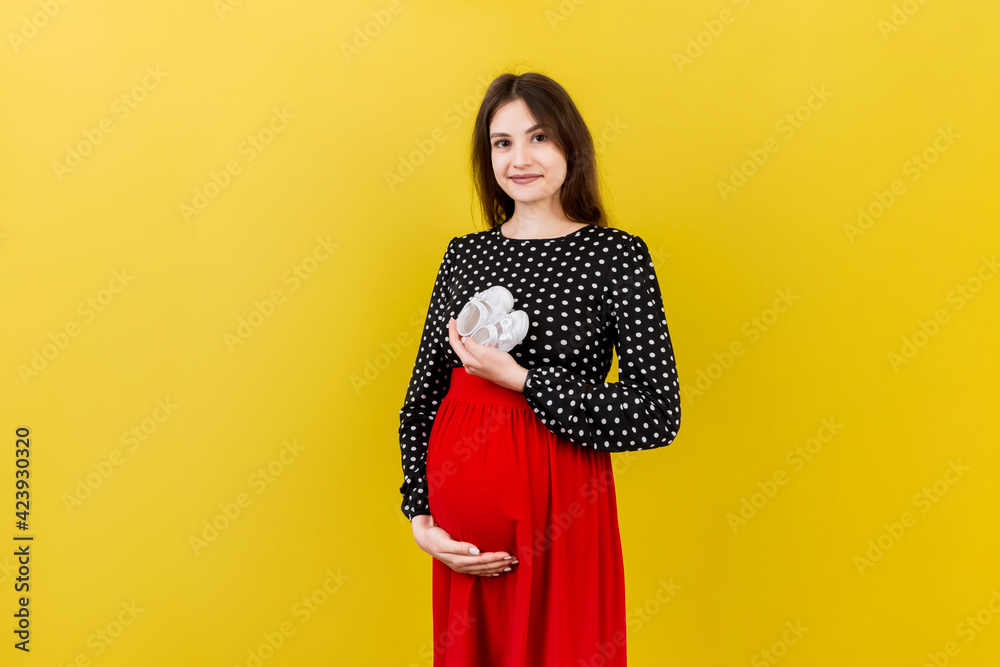 Happy pregnant woman holding baby girl booties against her belly at Colored background. Happy pregnancy. Copy space Happy pregnant woman holding baby girl booties against her belly at Colored background. Happy pregnancy. Copy space