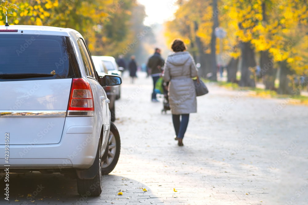Cars parked in a row on a city street side on bright autumn day with blurred people walking on pedestrian zone. Cars parked in a row on a city street side on bright autumn day with blurred people walking on pedestrian zone.