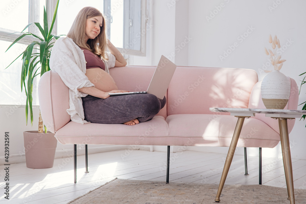 Young pregnant woman working on laptop at home in neutral interior. Working mother. Home office Young pregnant woman working on laptop at home in neutral interior. Working mother. Home office