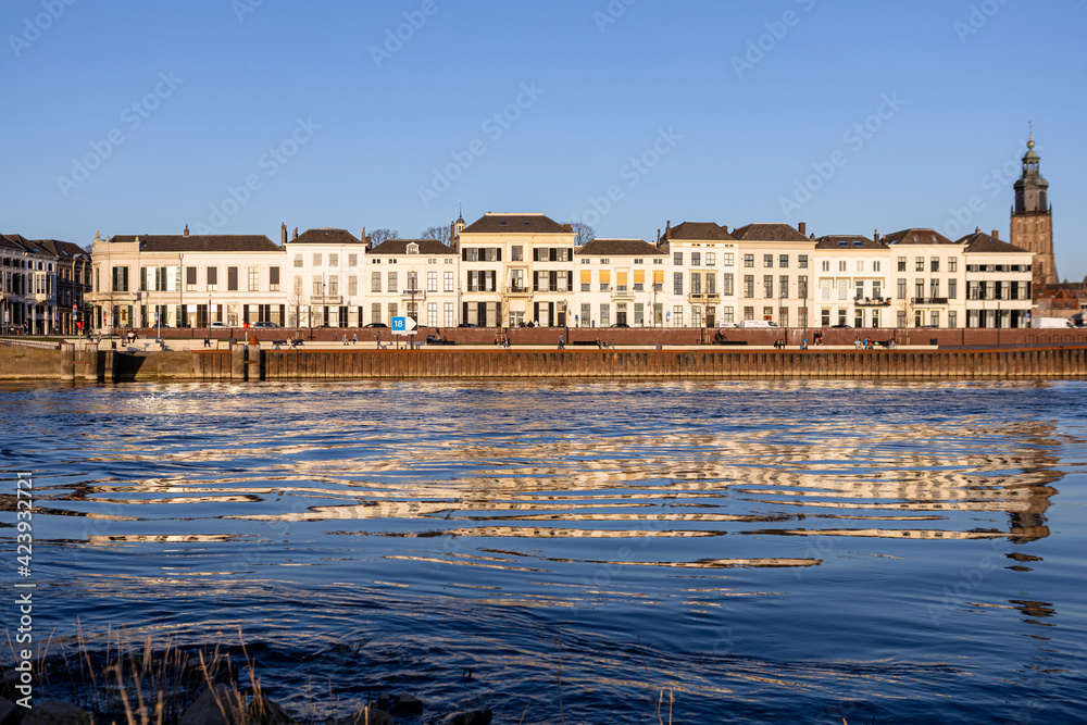 Cityscape countenance of historic Hanseatic Zutphen, The Netherlands, reflecting in the river IJssel seen from the opposite shore at sunset against a clear blue sky