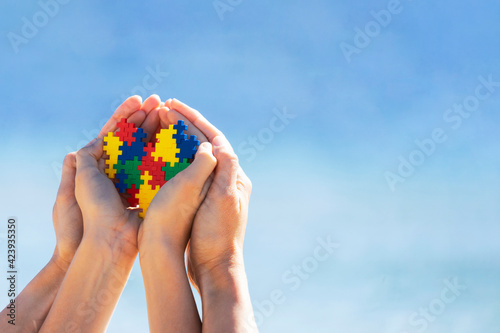 Fototapeta Autistic kid hands with multicolored puzzle heart supported by mother hands over blue sky background. World autism awareness day