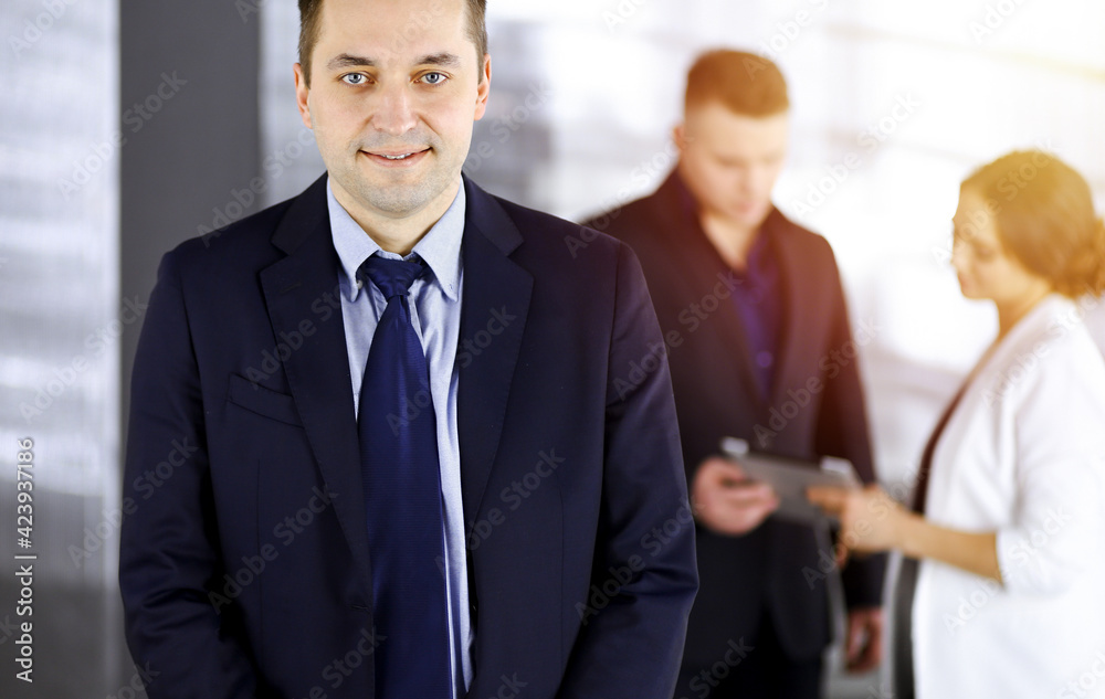 Fototapeta premium Portrait of a self-confident middle aged businessman in a blue suit, standing in a sunny modern office with his colleagues at the background. Concept of business success