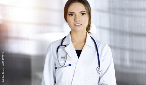Portrait of a professional friendly woman doctor with a stethoscope, standing straight in a clinic. Physician at work in a sunny hospital. Perfect medicine and healthcare concept
