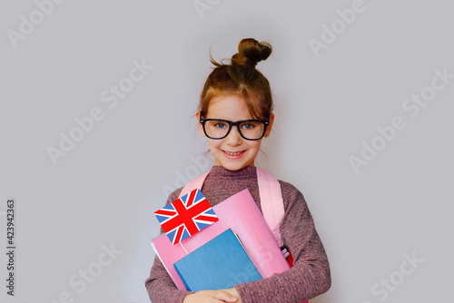 Happy girl kid 6 y.o. with black glasses holds a miniature of the English flag ,textbooks, and a school backpack. Learn English. Concept of education.
