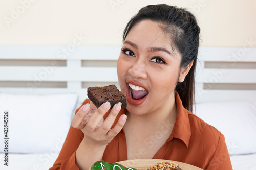 Happy obese female eating a tasty brownie in bedroom. Young plus size woman posing with brownie on bed at home. Overweight female sitting on bed with plate donuts. Unhealthy eating concept.