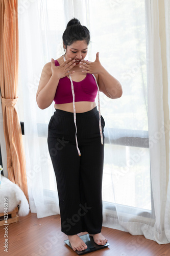Overweight woman standing on weight scale in bedroom. Obese woman with measure tape and weighing machine. Overweight woman standing on weighing machine. Surprise female on weighing machine at home.