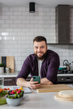Happy Young Man smiling and holding a Smartphone in the Kitchen with Vegetables on the Table online searching vegetarian recipe ideas on food app, Copy Space, Portrait