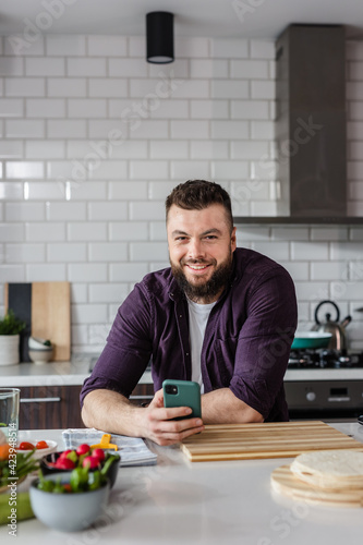 Happy Young Man smiling and holding a Smartphone in the Kitchen with Vegetables on the Table online searching vegetarian recipe ideas on food app  Copy Space  Portrait