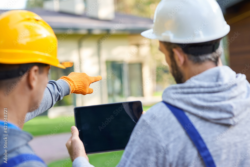 Use your vision. Close up of two young male builders using digital tablet pc, discussing ideas while working at cottage construction site Use your vision. Close up of two young male builders using digital tablet pc, discussing ideas while working at cottage construction site