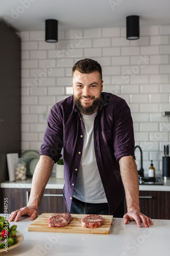 Young Man preparing Food Raw Steak Meat on a Wooden Board and Smiling at Camera at Home in Modern Kitchen, Ready to cook, Portrait, Copy Space