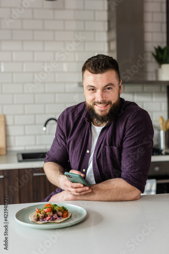 Happy young man preparing dinner using Smartphone App and smiling at Home in Modern Kitchen on a Sunny Day, Portrait