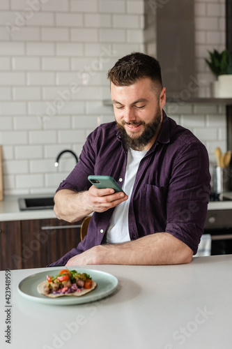Man taking photos with food using a smartphone at home in modern kitchen sitting at table ready to eat tasty meal with Mexican food on Daylight  Portrait 