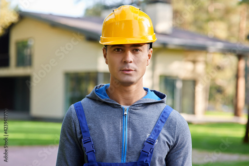 Portrait of handsome young male engineer in hard hat looking at camera, posing outdoors while working on cottage construction