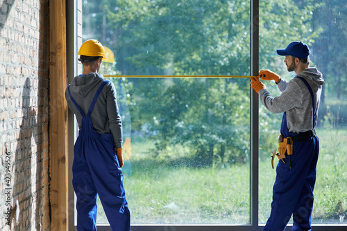 Making new home. Rear view of two young male builders wearing blue overalls using measuring tape while working on cottage construction site