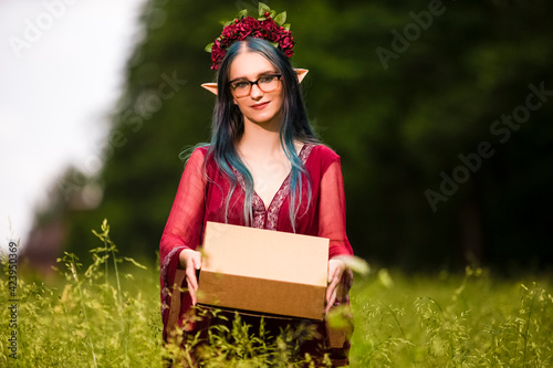 Portrait of Positive Caucasian Girl Posing in Flowery Garland and Artistic Ears Offering Gift Box on Green Field. photo