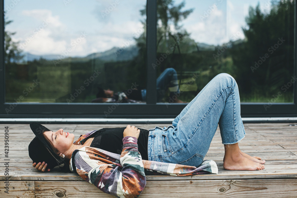 Stylish happy woman relaxing on wooden terrace at modern cabin with windows in mountains. Travel - obrazy, fototapety, plakaty 