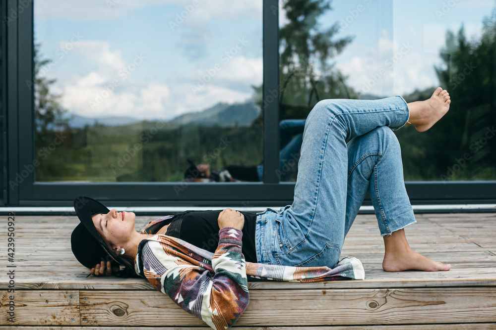 Stylish happy woman relaxing on wooden terrace on background of modern cabin in mountains. Tranquil - obrazy, fototapety, plakaty 