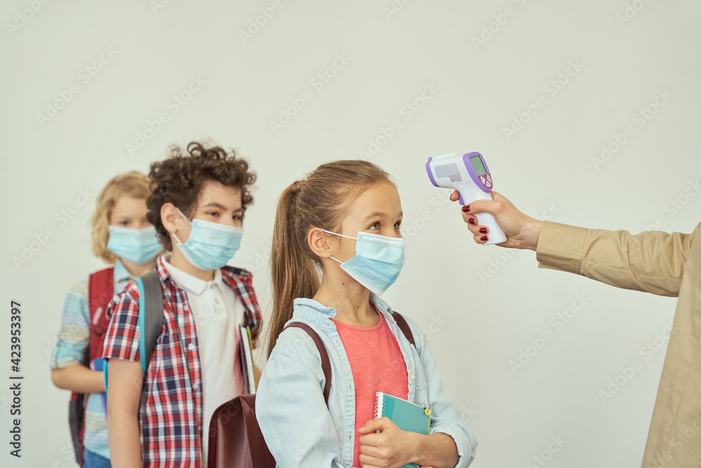 Little girl watching her teacher measuring temperature, screening kid with digital thermometer while students coming to school Little girl watching her teacher measuring temperature, screening kid with digital thermometer while students coming to school