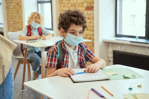 Social distancing. School boy wearing face mask writing in his notebook while sitting at the desk in a classroom. Kids studying in elementary school
