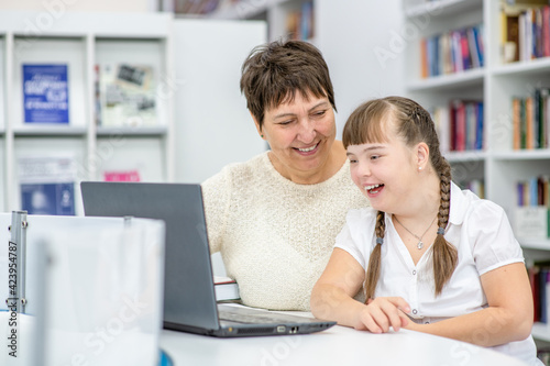 A girl with down syndrome is uses a laptop with her teacher at library. Education for disabled children concept © Ermolaev Alexandr