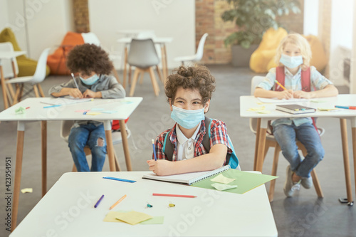 Happy school boy wearing face mask during coronavirus outbreak smiling while sitting at the desk in classroom. Kids studying in elementary school