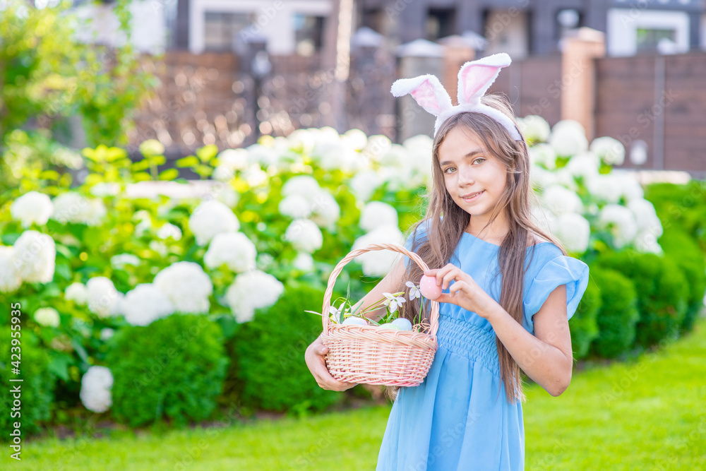 Fototapeta premium Smiling young girl wearing Easter bunny ears holds basket of a Easter eggs. Empty space for text
