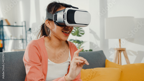 Asian lady wearing headset glasses of virtual reality gesticulating hand sitting on couch in living room at house. Stay at home covid quarantine, Re-imagining Reality, VR technology of future concept.