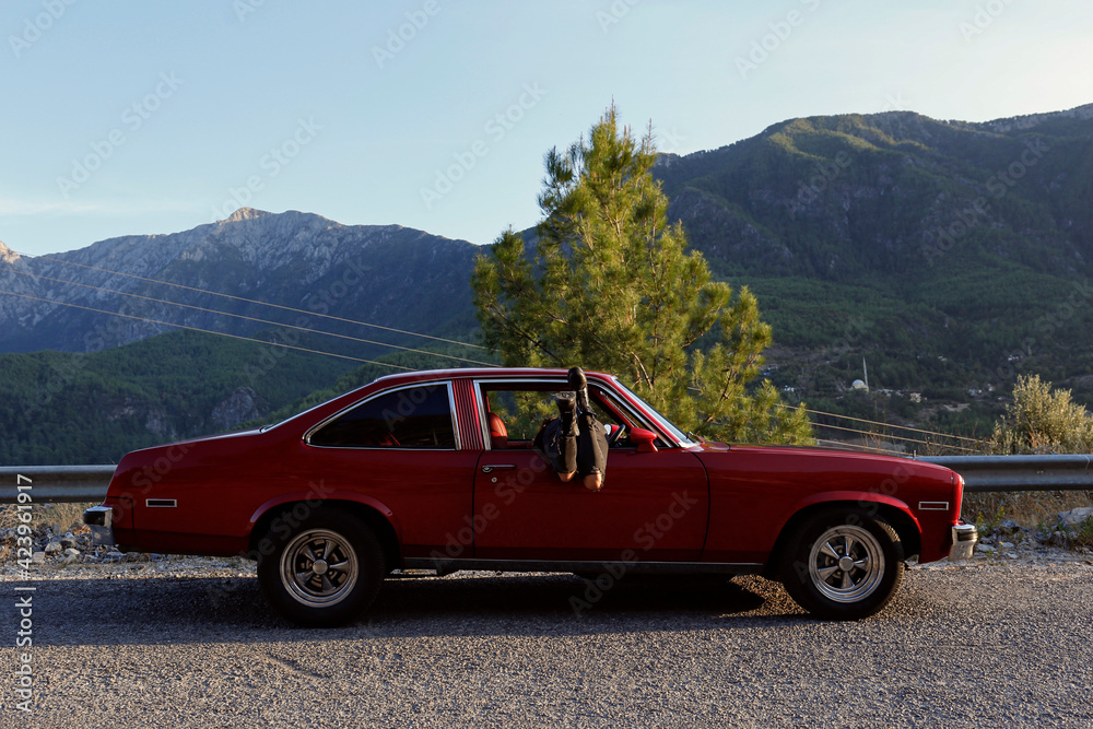 Young carefree woman wearing black ripped jeans and moto boots with her legs sticking out of th window of cherry red muscle car. Traveling alone concept. Copy space, background, mountain view.