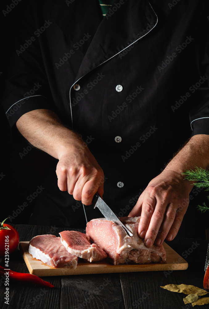 Chef Butcher cutting pork meat with knife on kitchen, cooking food. Vegetables and spices on the kitchen table to prepare delicious lunch