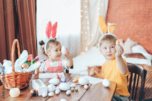Preparing for Easter  children paint eggs for religious holiday. Boy and girl are painting Easter eggs for the holiday at wooden table. Children have bunny ears on their heads