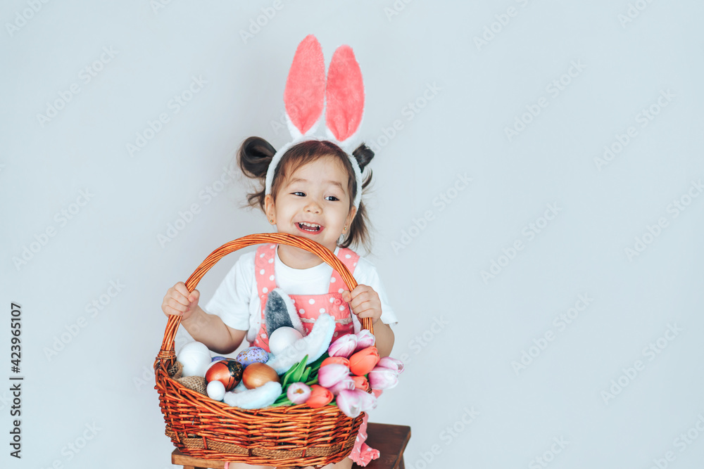 Little girl dressed as rabbit holding large basket filled with eggs and tulips against white background