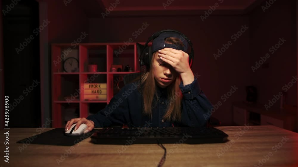Upset girl gamer sits at night at the computer and shows a gesture of facepalm, lowered his eyes down from the shame of losing the game
