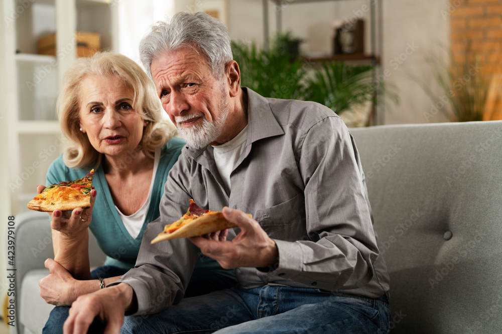 Cheerful husband and wife sitting on sofa at home. Happy senior woman and man eating pizza while watching a movie