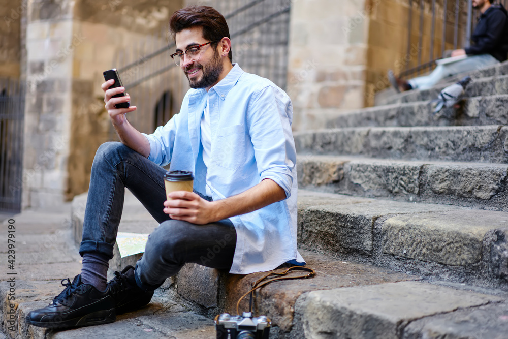 Content young man in eyeglasses using smartphone while sitting on stairs of old building