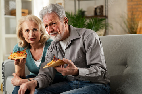 Cheerful husband and wife sitting on sofa at home. Happy senior woman and man eating pizza while watching a movie