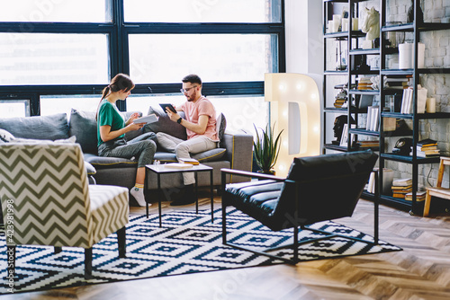 Caucasian male resting at comfortable sofa with wife searching best seller on touch pad website during hobby time in flat, boyfriend prefer electronic ebook why girlfriend reading paper literature