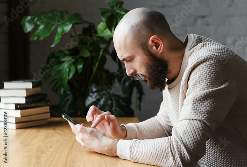 bald man 35 years old with a beard and mustache in a beige jumper sits at a table with a phone