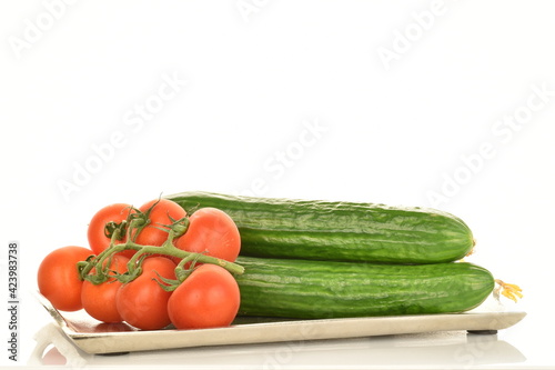 Several green cucumbers and red ripe, cocktail tomatoes on a branch with a metal tray, close-up, isolated on white.
