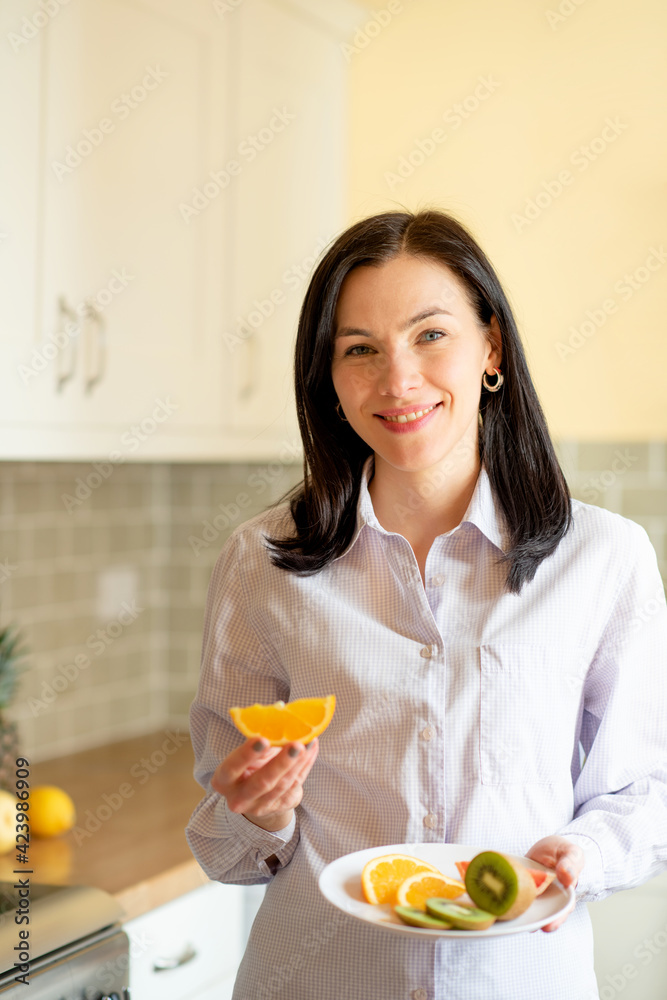 A smiling woman in white shirt holding fruits in a kitchen