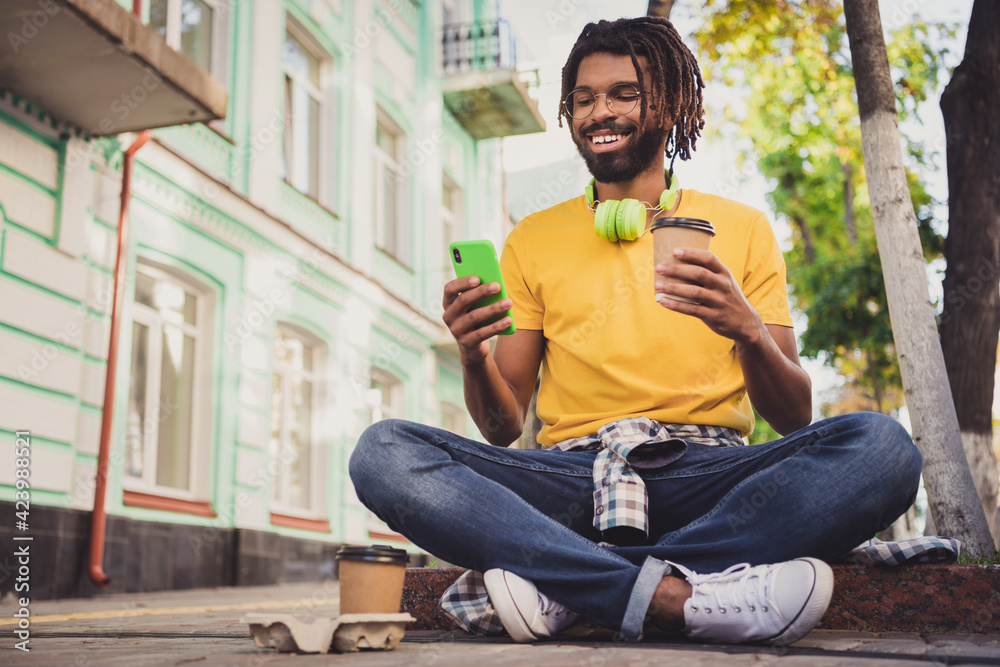 Photo portrait of young guy sitting on the ground reading news from smartphone drinking beverage wearing earphones spectacles - obrazy, fototapety, plakaty 