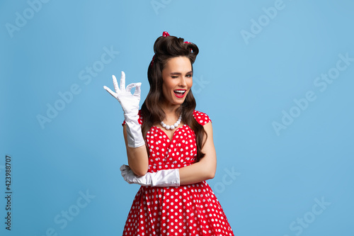 Beautiful young pin up woman in polka dot red dress and gloves showing ok gesture and winking on blue background