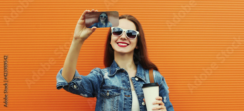 Portrait close up of smiling woman taking a selfie picture by smartphone wearing a denim jacket on an orange background