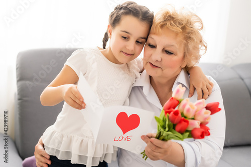 Sincere cute little granddaughter strong cuddling and kissing in cheek her 60s grandmother gave her pretty spring flowers congratulates with birthday, International Womens day, close up concept image
