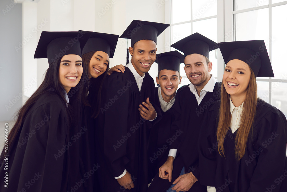 Portrait of a group of young and cheerful students of different nationalities posing by the window in a bright university classroom. Concept of education, graduation and alumni.