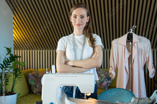 Young woman dressmaker poses in a workshop, arms crossed against the background of a sewing machine