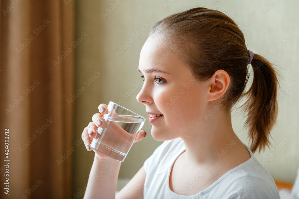 Happy teen girl maintains water balance for body health by drinking transparent cup of clean water. Smile young woman drinks glass of pure water in morning after waking up Happy teen girl maintains water balance for body health by drinking transparent cup of clean water. Smile young woman drinks glass of pure water in morning after waking up